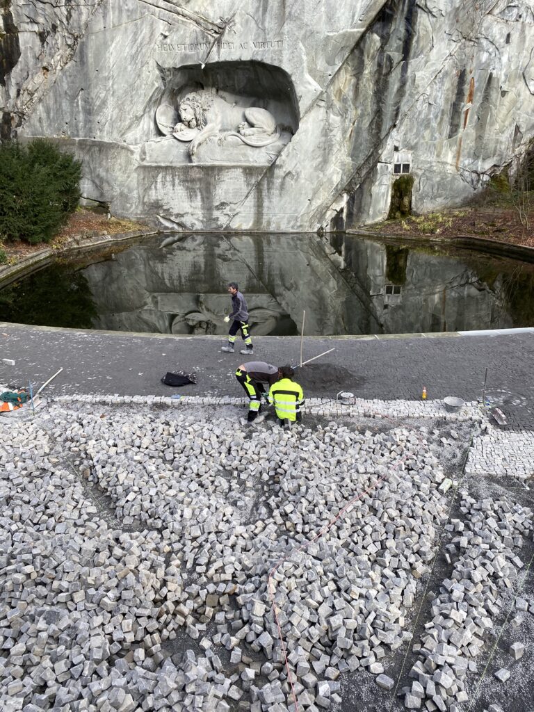 Vorne im Bild liegen die Pflastersteine, die noch versetzt werden. Entlang des Wassers ist die fertige Pflästerung bereits mit Fugensand bedeckt. Löwendenkmal in Luzern.